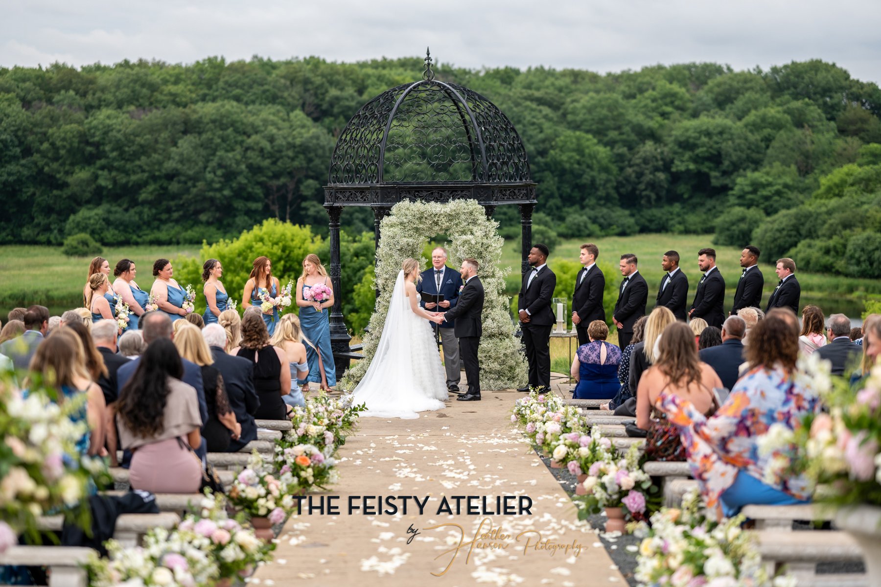 An elegant outdoor wedding ceremony with the aisles lined with pastel flowers and a giant arch, bride and groom centered with their bridal party and guests surrounding them.