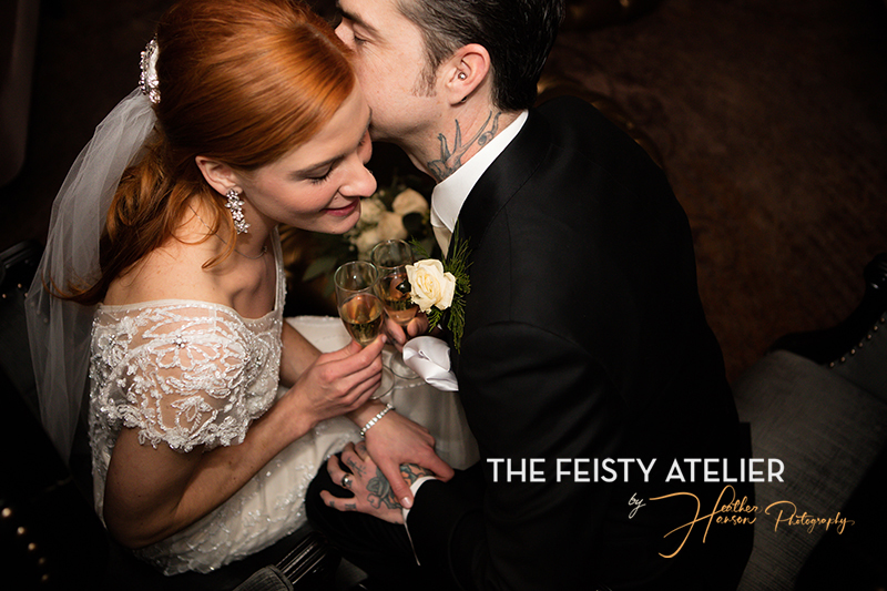 A bride leans in and listens intently as her groom whispers something in her ear. She hols a champagne glass and smiles.