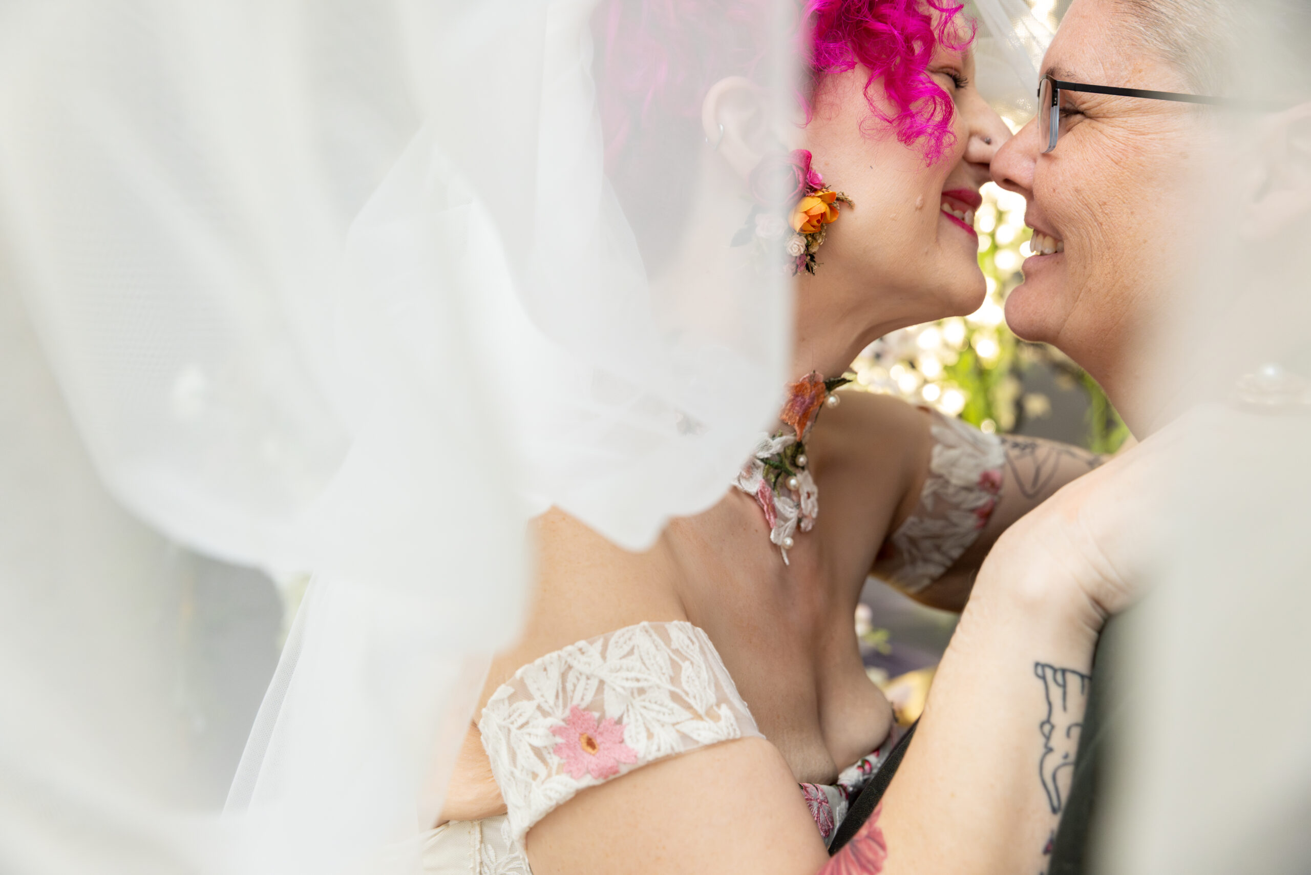 Two brides embrace and smile at each other underneath a veil.