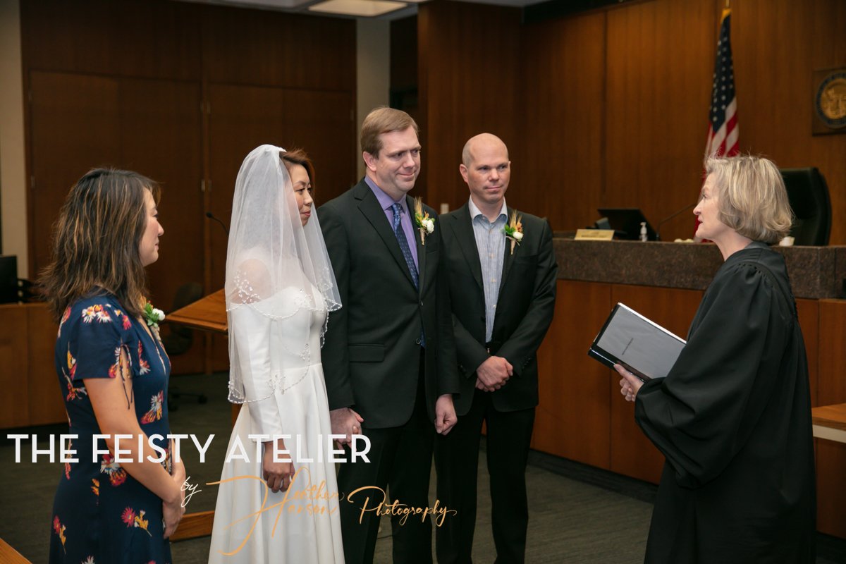 A bride and groom stand, surrounded by two witnesses in front of a judge as she marries them inside a courthouse.
